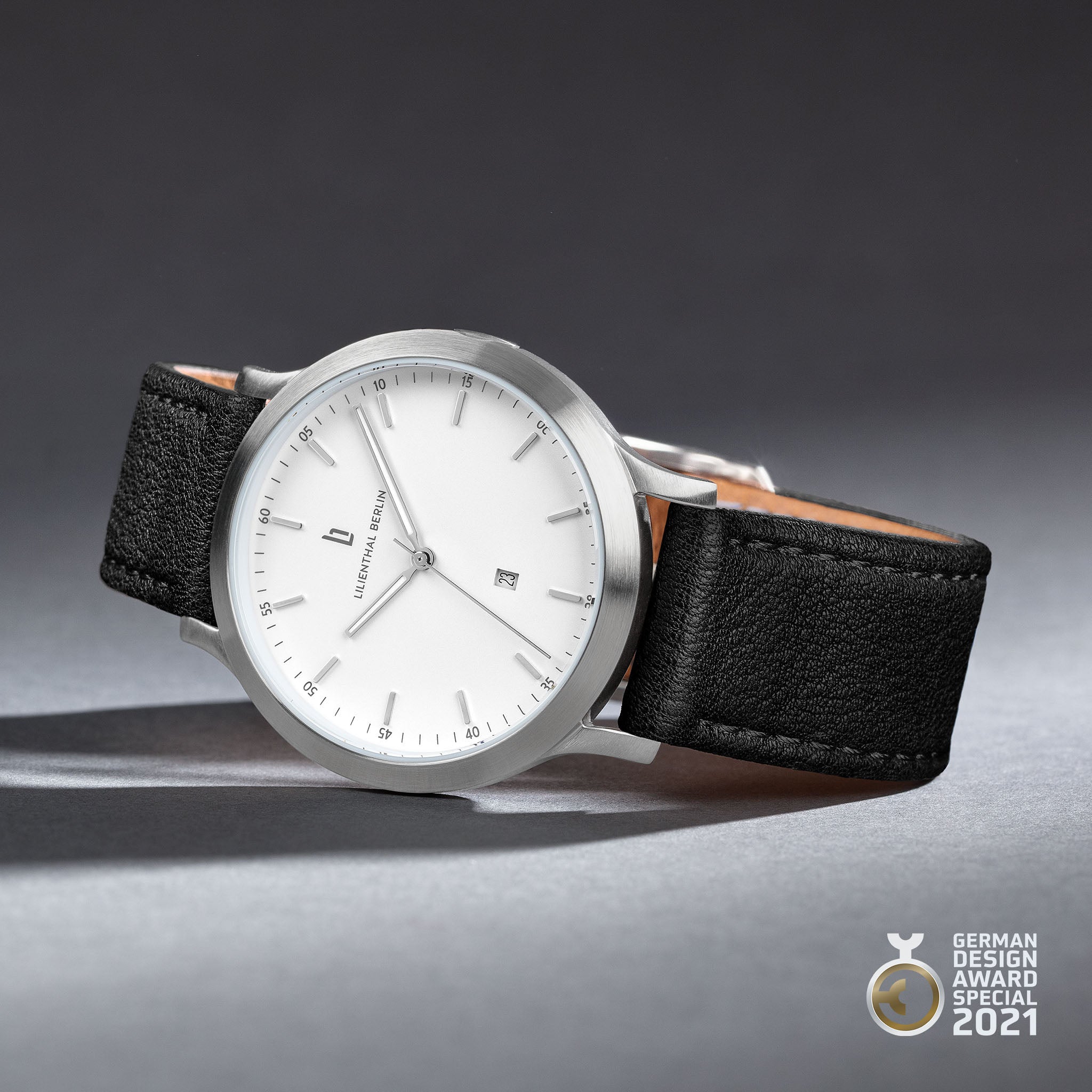 Minimalist silver watch with white dial, black leather strap, date window, award logo.