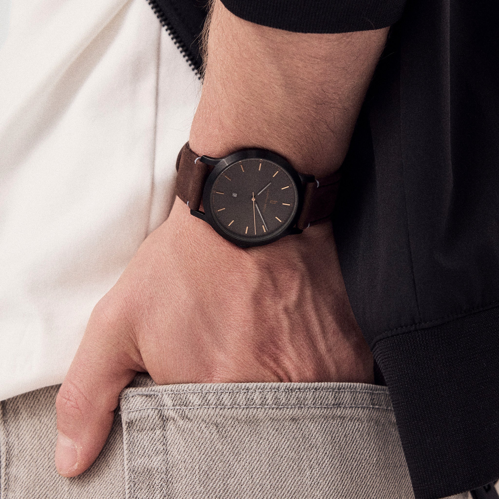 Minimalist wristwatch with dark face and brown leather strap on man's wrist in casual outfit