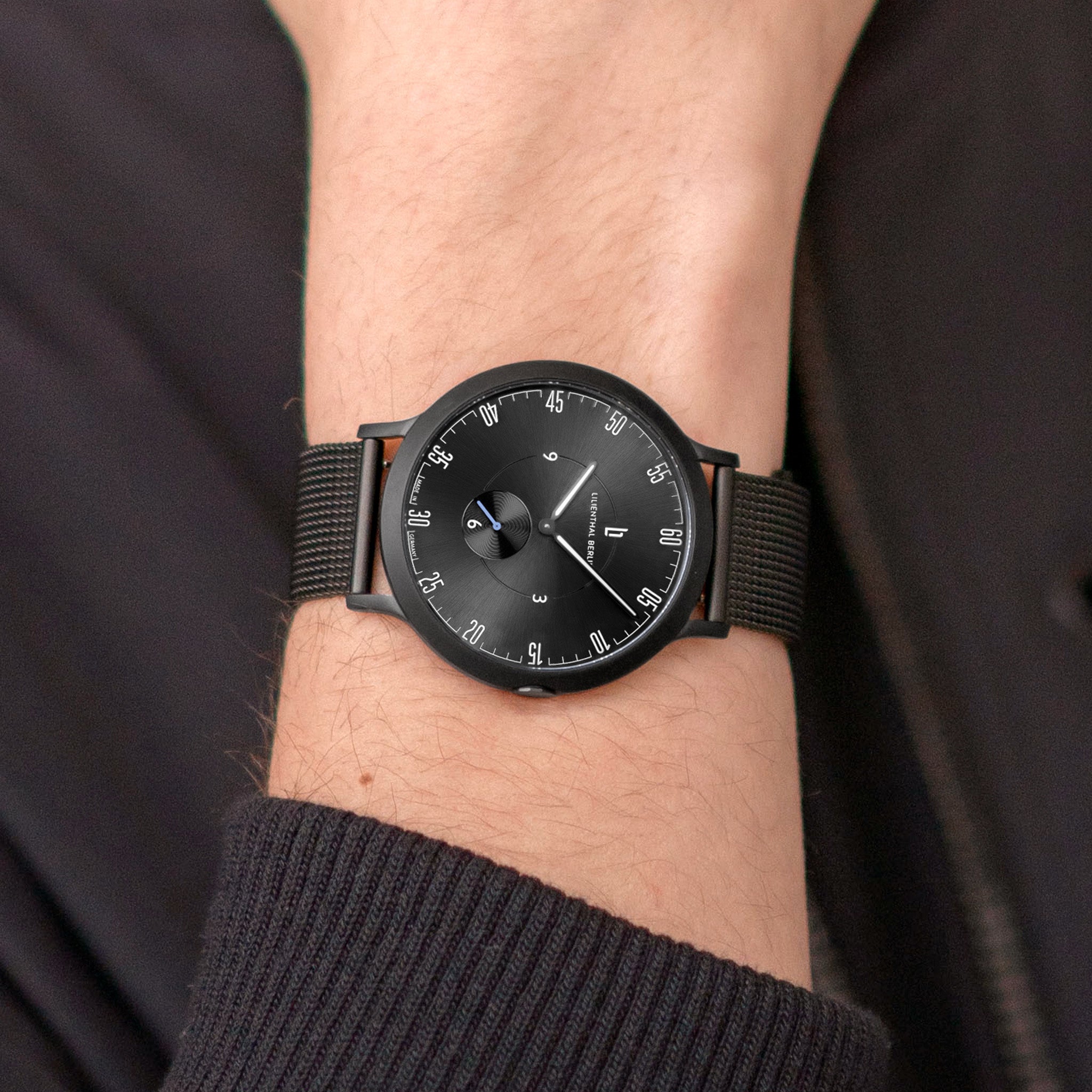 Minimalist black wristwatch with mesh strap on wrist, modern dial design, close-up view