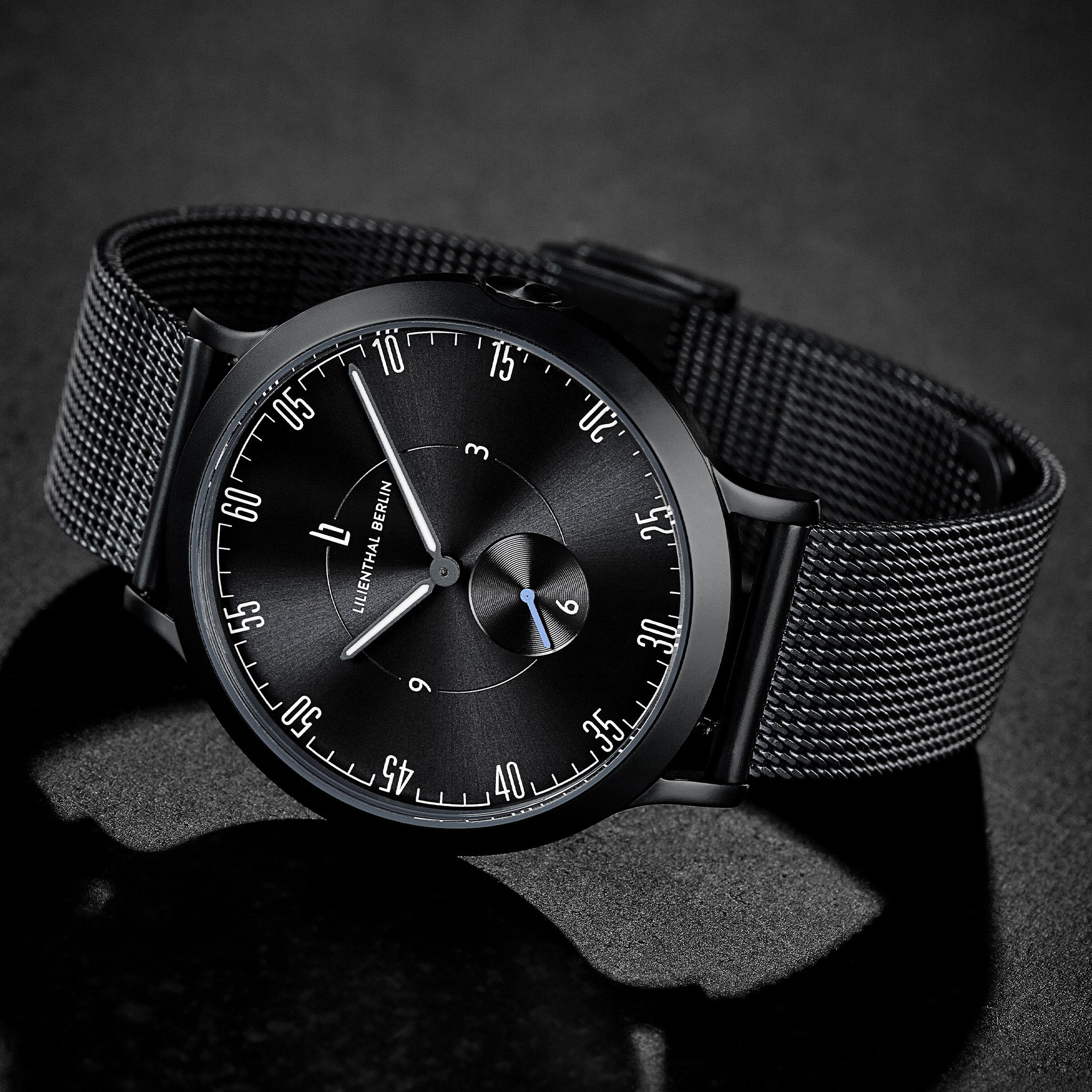 Minimalist black mesh strap wristwatch with black dial on a dark background