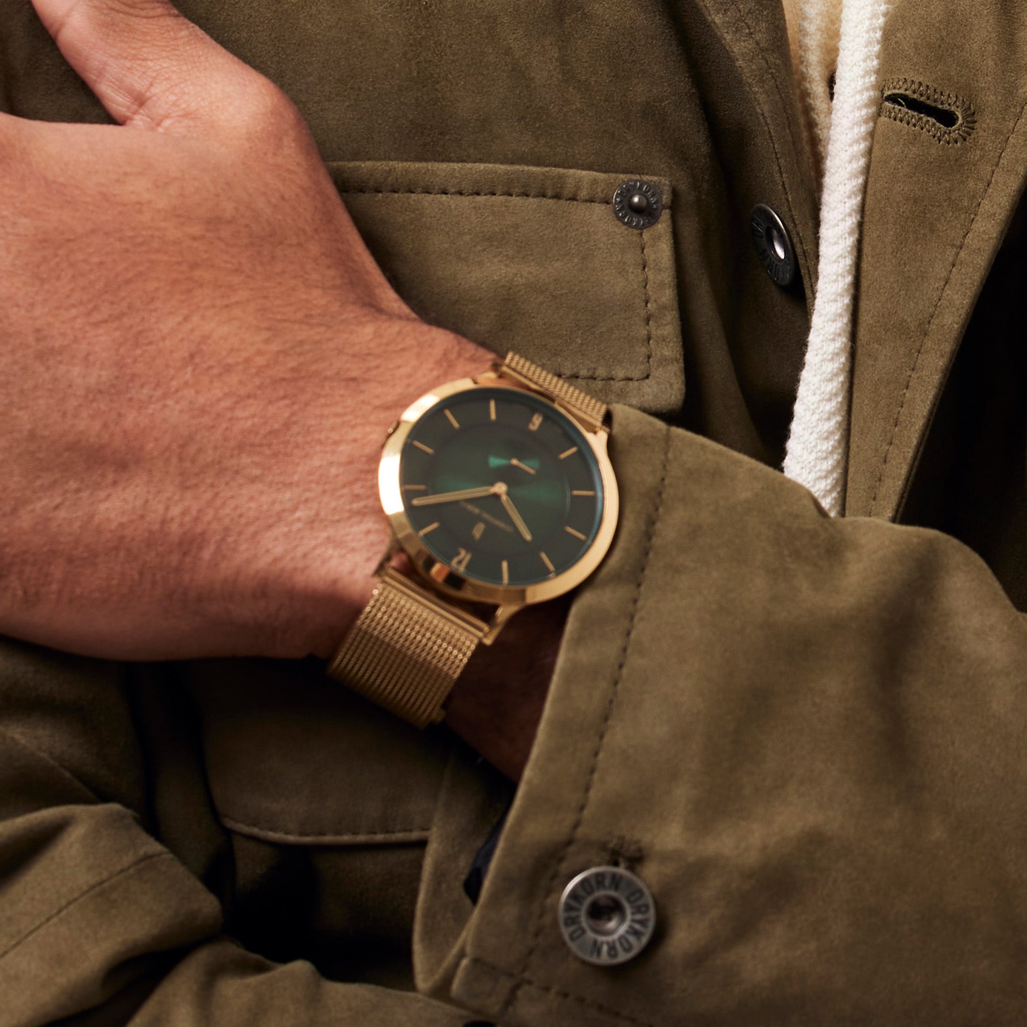 Gold mesh wristwatch with green dial on wrist, styled with green jacket