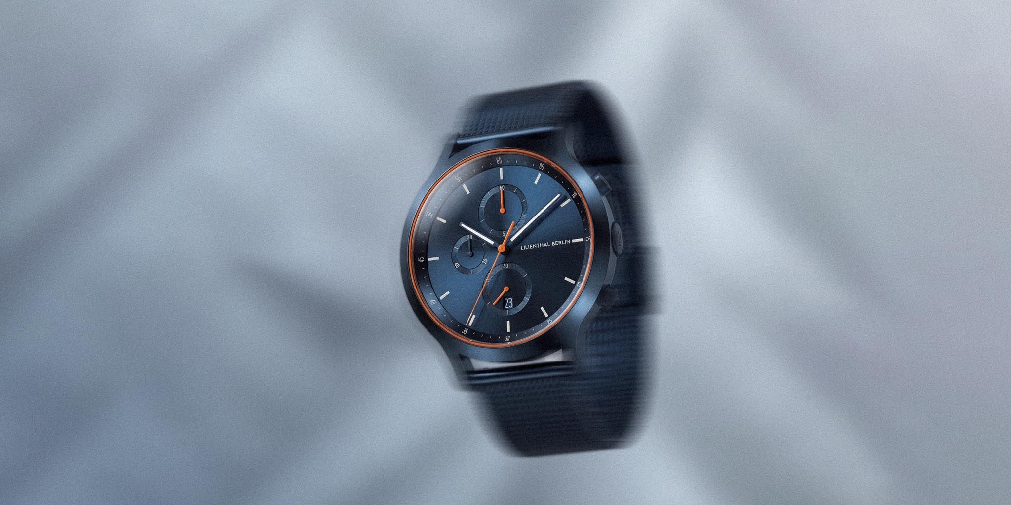 Blue chronograph watch with orange accents and mesh strap on a soft grey background