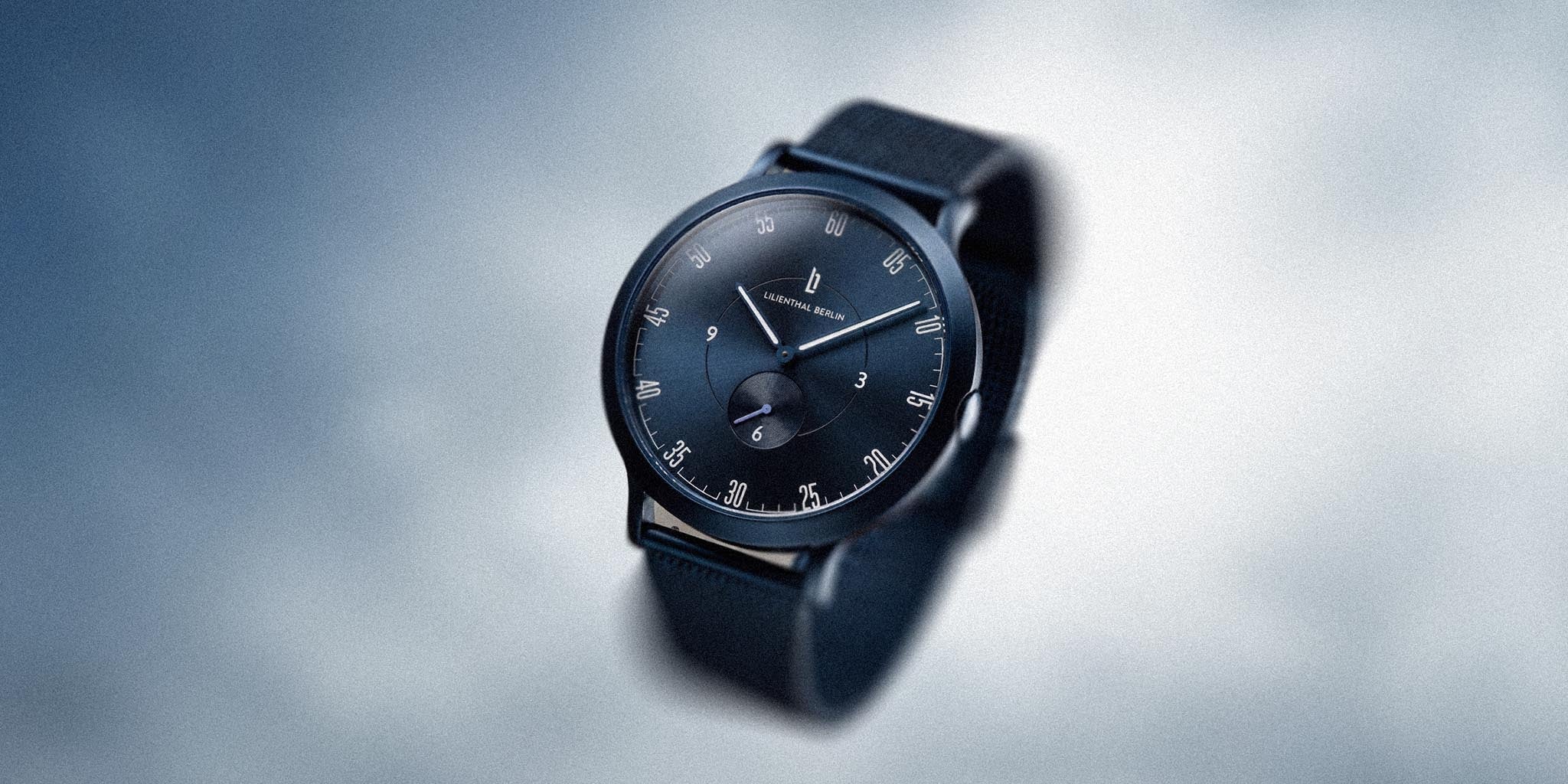 Minimalist blue wristwatch with black strap and silver details on a soft cloudy background