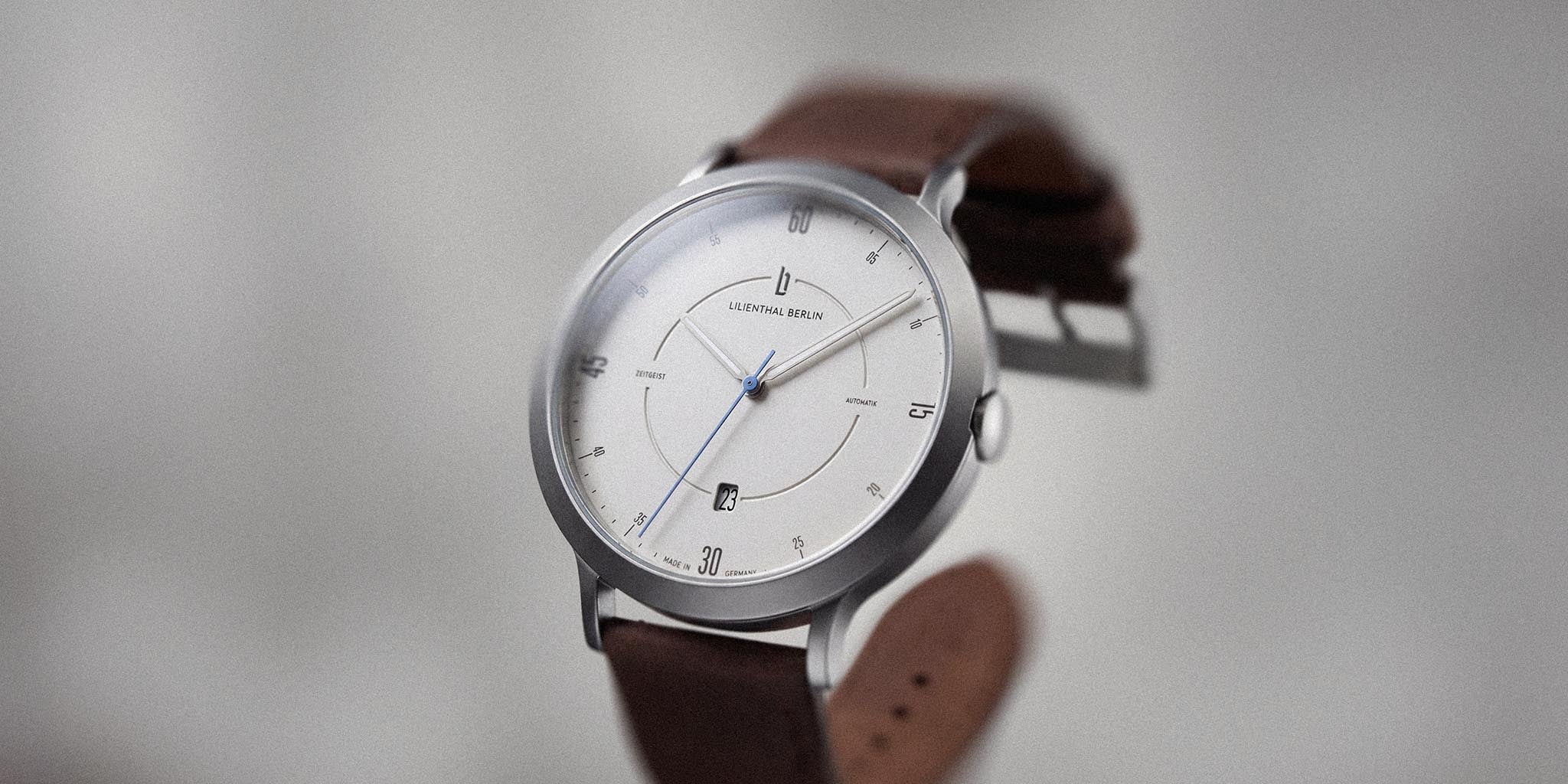 Minimalist silver and white automatic wristwatch with brown leather strap on neutral background