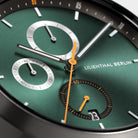 Close-up of a green chronograph watch face with subdials, orange hands, and date display.
