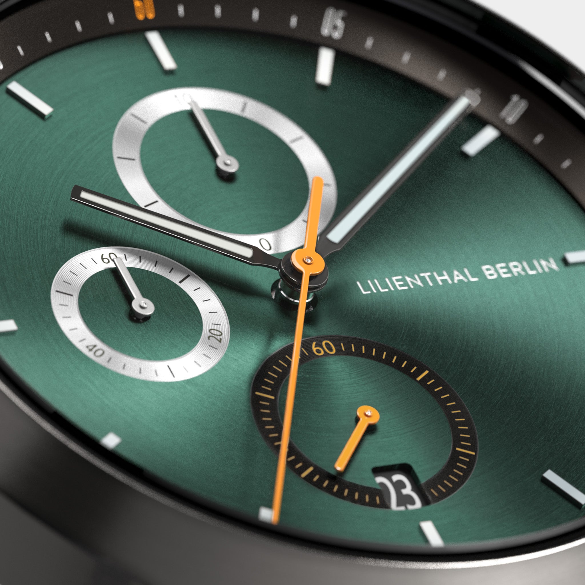 Close-up of a green chronograph watch face with subdials, orange hands, and date display.