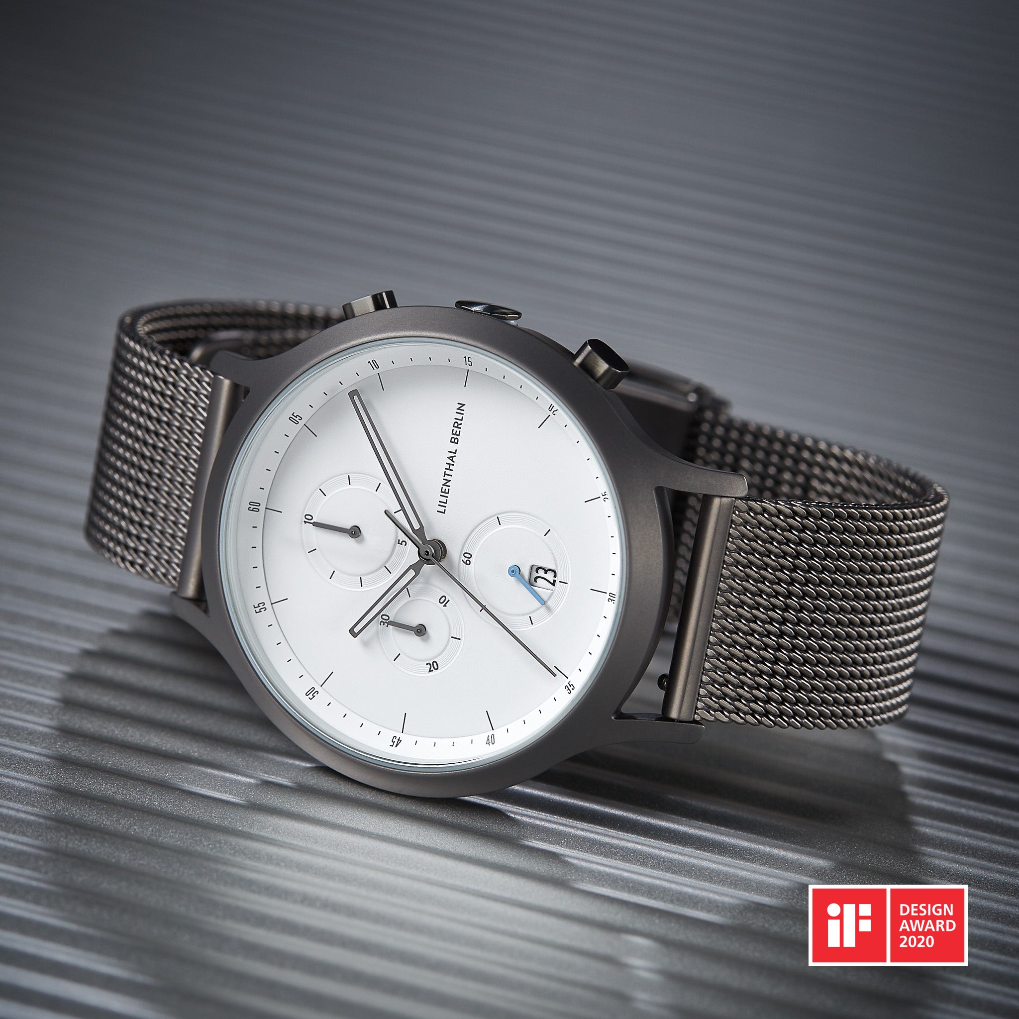Minimalist wristwatch with white dial, mesh band, and chronograph features on metallic surface