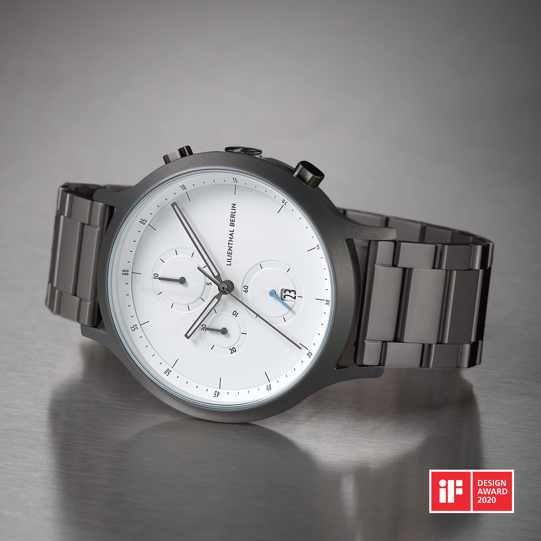 Minimalist silver chronograph watch with white dial, steel band, and date display on metal surface.