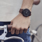 Man wearing a minimalist black watch with brown leather strap, hand on bicycle handlebar.