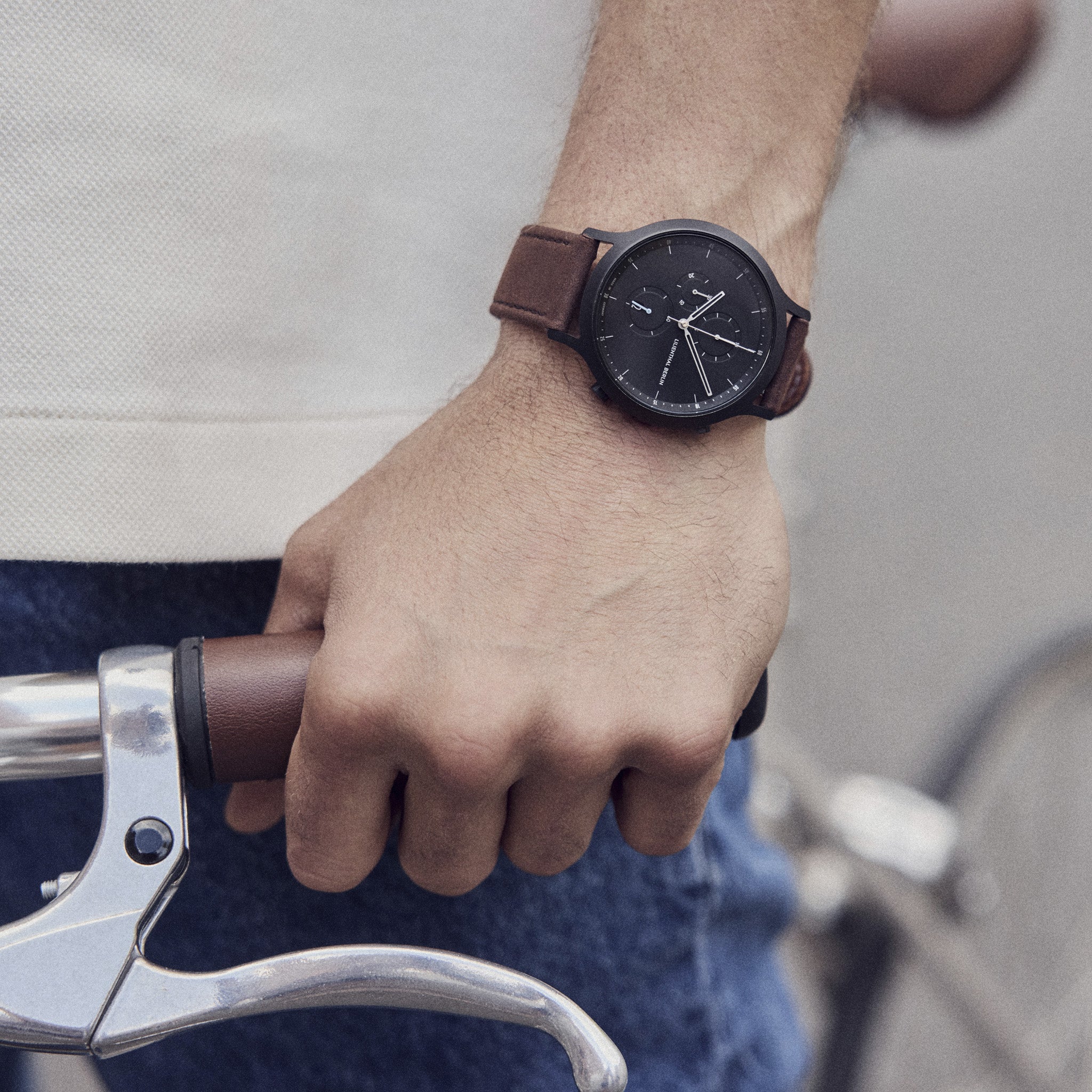 Man wearing a minimalist black watch with brown leather strap, hand on bicycle handlebar.