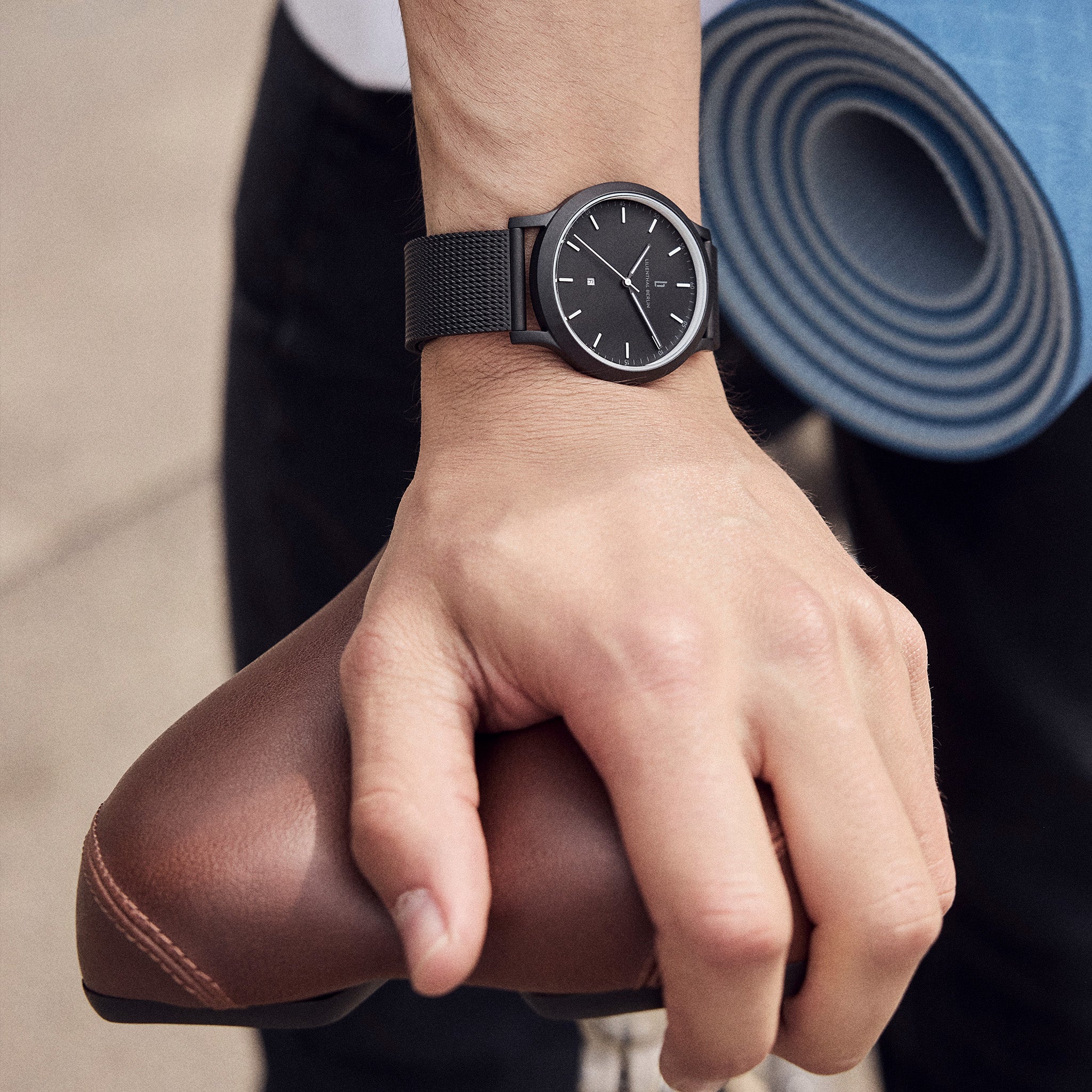 Minimalist black wristwatch with mesh band on wrist, hand holding brown bike saddle, yoga mat