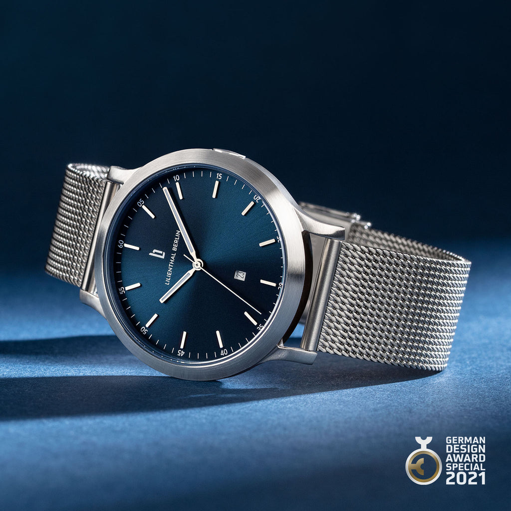 Minimalist silver mesh wristwatch with blue dial, date display, and German Design Award badge.