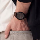 Minimalist black wristwatch with a brown leather strap on a person’s wrist in casual attire