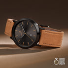 Minimalist black watch with brown leather strap, gold markers, and date display on beige background