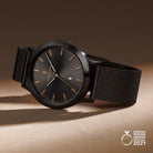 Minimalist black wristwatch with mesh strap, gold markers, date display, award emblem, studio lighting