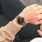 Minimalist bronze wristwatch with black dial and mesh strap on a person's wrist