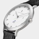 Minimalist silver analog watch with white dial, black leather strap, and blue second hand