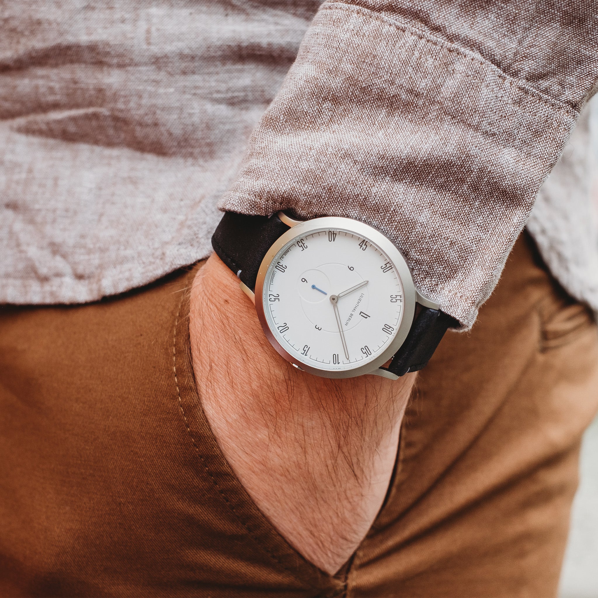 Minimalist silver wristwatch with white dial and black strap on wrist, hand in pocket