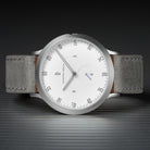 Minimalist silver wristwatch with white dial and grey suede strap on a dark striped surface