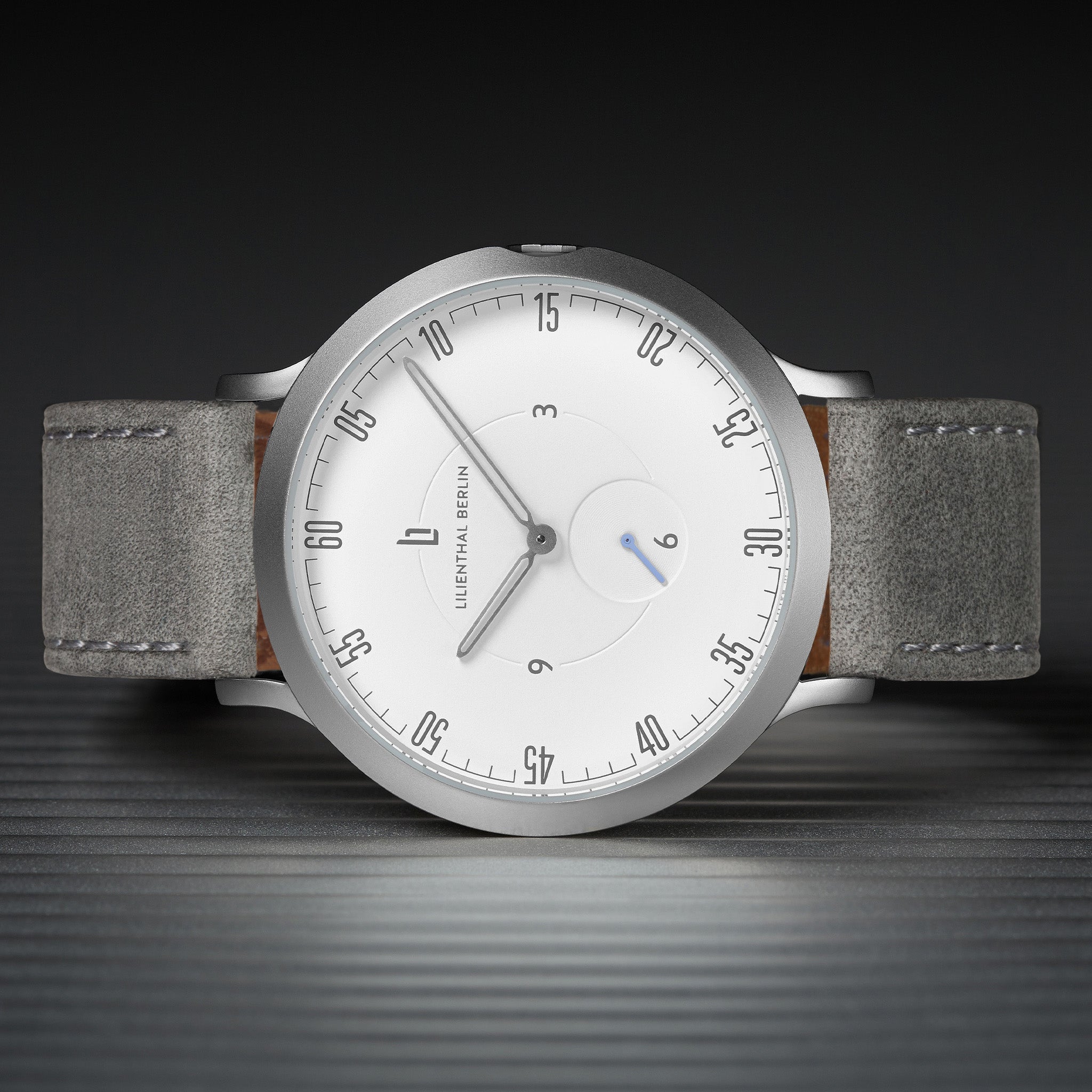Minimalist silver wristwatch with white dial and grey suede strap on a dark striped surface