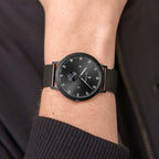 Minimalist black wristwatch with mesh strap on person's wrist, modern analog dial design.
