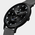 Minimalist black watch with black dial, white markers, and grey suede strap, close-up view.