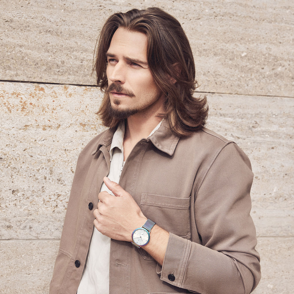 Man wearing a beige jacket and blue wristwatch standing against a stone wall