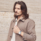 Man wearing a beige jacket and blue wristwatch standing against a stone wall