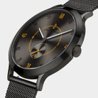 Close-up of a modern black mesh strap watch with a dark grey dial and yellow accents.