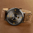Modern minimalist watch with brown textured strap, black dial, and orange accents on numbers